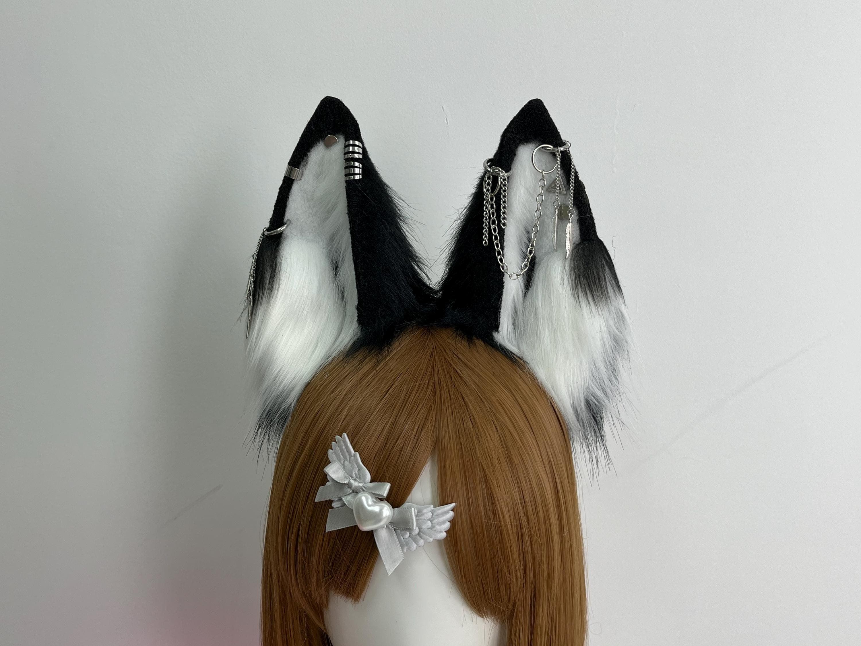 Detailed close up front view of black and white wolf ears showing intricate silver chain ear cuff piercings with feather drop charms and bar cuff accents, handcrafted faux fur construction revealing realistic wolf proportions fluffy white inner plush jet black outer fur. Ideal for gothic punk alt fashion therian cosplay anime kemonomimi character builds furry convention Halloween costume. Unique statement piece rave festival streetwear photoshoot prop gift for wolf lover.