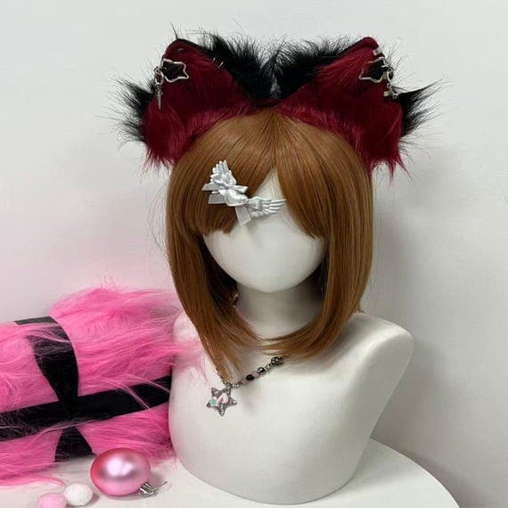 May include: A mannequin head with a brown wig, featuring red and black cat ears. A silver necklace with a star pendant and a white bow-shaped accessory are also visible. A pink fluffy item and a pink bauble are in the foreground.