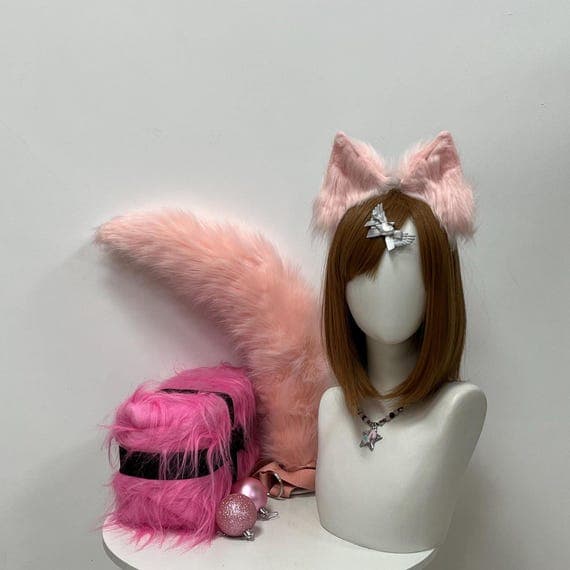 May include: A pink faux fur cat ear headband, tail, and a cube-shaped accessory with black bands. A mannequin head wears a brown wig, a silver bow, and a star pendant necklace. Decorative pink glitter balls are also present.